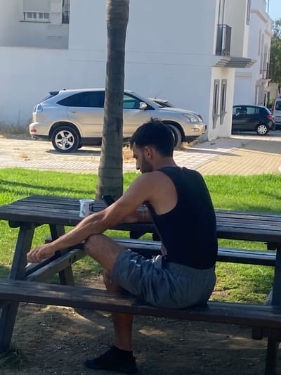 In Cancelada, Spain, a man sits at a wooden picnic table outdoors, facing away from the camera. He is wearing a black tank top and grey shorts. His left arm rests on the table, and his right leg is bent, with his foot resting on the ground. A white mug with a logo is on the table to his left. Behind him, a palm tree trunk is visible. In the background, a silver SUV is parked in front of a white building with balconies and windows. Further to the right, a black hatchback is also parked. The ground is a mix of grass and dirt.