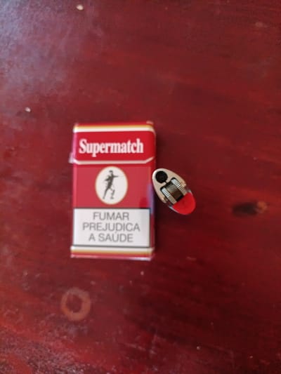 A pack of Supermatch cigarettes and a red lighter are placed on a reddish-brown wooden surface. The cigarette pack is red with white text that reads "Supermatch". Below the brand name, there is a white circle with a black silhouette of a person running. Further down on the pack, there is text in Portuguese that reads "FUMAR PREJUDICA A SAÚDE", which translates to "SMOKING HARMS HEALTH". The lighter is oval-shaped with a red base and a silver top. The image appears to be taken indoors, on a table, likely in a home or a casual setting, and not specifically in the city of Saurimo, Angola.