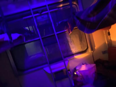 The photo appears to be taken inside a train compartment. A window with bars is visible. A striped blanket or shirt hangs over the top right of the window. Below the window, there’s a bag and some dark clothing. The lighting is a mix of blue and orange hues. The overall scene is somewhat blurry and disorienting.