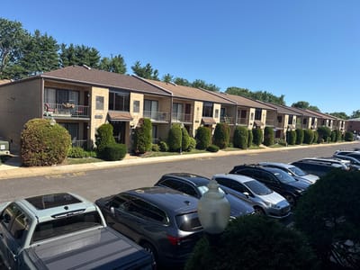 The image shows a row of tan brick apartment buildings under a clear blue sky. The buildings have brown roofs and dark brown trim around the windows and doors. Each unit appears to have a balcony on the second floor. Numbered signs like "C25-28" are visible on the building facades. In front of the buildings is a paved parking lot with several cars parked in a line. A "FIRE LANE" marking is visible in yellow paint on the asphalt. Green trees and manicured bushes are planted along the front of the buildings and bordering the parking lot. The scene is devoid of people and appears to be a residential area. There is no identifiable information suggesting the city of Philadelphia or the United States of America.
