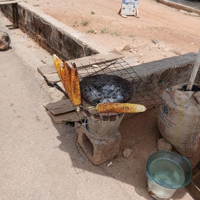 The image shows a street vendor in Kufang, Nigeria, grilling corn on the cob over a charcoal brazier. There are four ears of corn, three of which are partially grilled. The corn is arranged vertically, with the top of the corn closest to the camera. There is a metal grate over the brazier, holding the corn in place. There is a metal pot beneath the grate to catch any drips. There is also a large bag of cement on the ground, a bowl of water, and a wooden board to the left of the brazier. The scene is set on the side of a street, with a concrete gutter to the left of the brazier. The ground is dusty and dry. There are no people present in the scene.