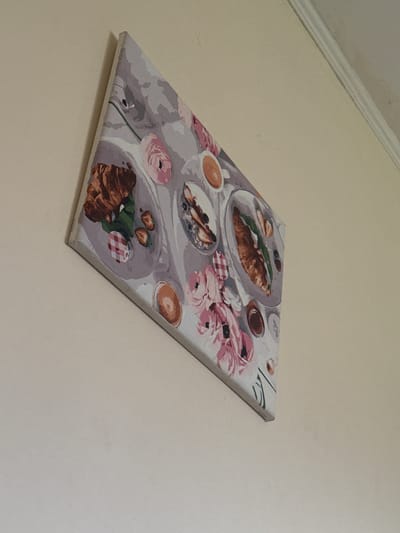 A framed canvas print is displayed on a light-colored wall. The print depicts a top-down view of a breakfast spread, featuring pastries, fruit, and cups of coffee. Pink flowers are also visible within the artwork. The print is hung at an angle on the wall, which appears to be in a residential setting, possibly in Horenychi, Ukraine, given the information provided.