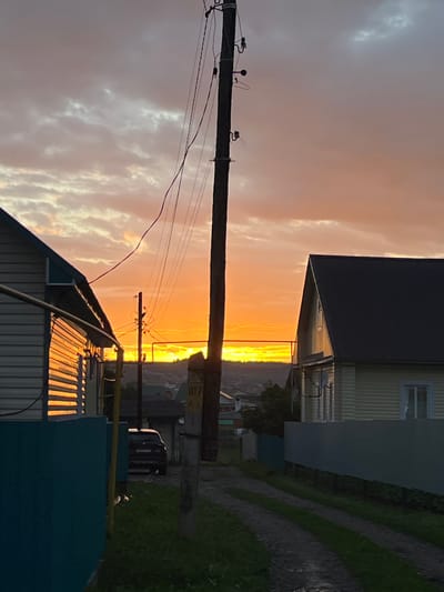 The image depicts a sunset over a rural village, likely located in Большой Кукмор, Russia. The sky is filled with vibrant orange and pink hues, with scattered clouds catching the light. A wooden utility pole with electrical wires is prominently positioned in the foreground, dividing the scene. On the left, the side of a light-colored house with horizontal siding is visible, reflecting the sunset's glow. To the right, another house with a dark roof and light-colored siding stands. A dark-colored car is parked on a dirt path between the houses. The ground is covered with grass and a dirt path, suggesting a residential area. The overall impression is a peaceful, rural evening scene.