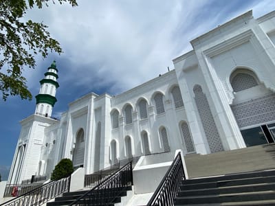 The image displays a white mosque with a green-topped minaret, located in Aluekambuk, Indonesia. The mosque features traditional Islamic architectural elements, including arched windows with intricate geometric patterns. A set of stairs with black railings leads up to the entrance of the mosque. Green trees and branches are visible in the upper left corner of the frame, contrasting with the white structure and blue sky. The weather appears partly cloudy.