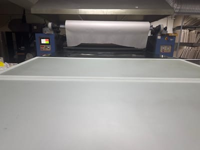 In Bahçelievler, Turkey, a large white roll of paper is mounted on a blue machine, suggesting an industrial or printing environment. The machine has a digital display showing various parameters, including "5.0m/d," "210-," and "757.5." There are also buttons and indicators on the machine, typical of a piece of manufacturing equipment. To the right of the machine, shelves filled with what appear to be large, flat items, possibly paper or artwork, are visible. The overall setting is an indoor space with industrial-style lighting and ventilation visible on the ceiling. The foreground is occupied by a large, flat, white surface, potentially a table or another part of the machinery.