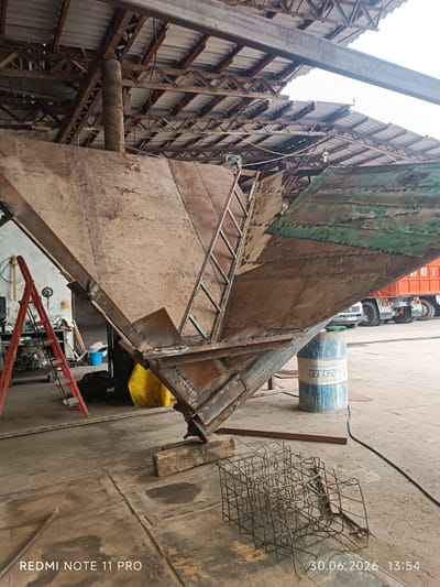 The image depicts a large, unfinished metal structure, likely a hopper or part of a large industrial machine, inside a workshop or storage area. The structure is made of rusted metal plates, with visible welding seams and some sections painted green. A metal ladder is attached to the side of the structure, suggesting it's designed to be accessed from multiple points or is in a stage of construction where it needs internal access.
In the foreground, there is a blue and white barrel with "FOSSER" printed on it, along with a metal cage-like object made of rebar. To the left, a red stepladder is leaning against a wall, and some tools and equipment are visible in the background, including what appears to be a generator. Further back, the rear end of an orange truck is visible, suggesting the workshop is located near a road or transport hub. The ceiling of the workshop is constructed with corrugated metal roofing supported by a metal framework.
The scene is indoors, in what appears to be a functional but somewhat disorganized workspace. The lighting suggests it is daytime, but the exact time of day is difficult to determine. There are no people visible in the image. The overall impression is one of ongoing construction or repair work.
The text visible at the bottom of the image reads "REDMI NOTE 11 PRO" and "30.06.2026 13:54".
