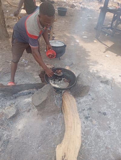 A young man, wearing a red and white striped shirt, is cooking over an open fire in Assai Sabongida, Nigeria. He is stirring food in a large black pot with a long wooden spoon. The pot is sitting on top of a large wooden log that is resting on a pile of rocks. There is a smaller pot to the right of the man. The man is standing on a dusty ground, and there is a tree trunk behind him. In the background, there is a wooden table and a partially visible person. The scene is a typical example of outdoor cooking in a rural setting.