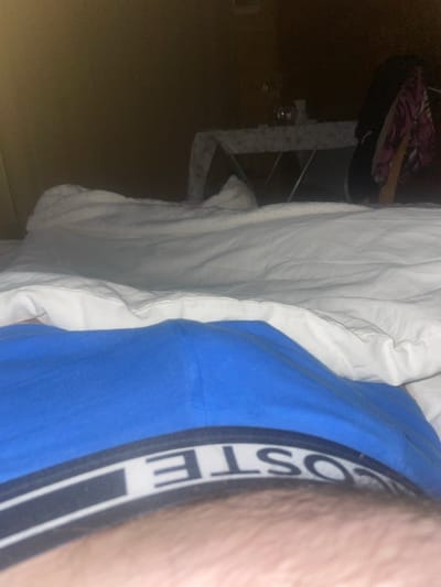 In a dimly lit room, likely a bedroom, a person is lying in bed covered by white linens. The lower portion of the image shows the waistband of blue underwear with the logo "LACOSTE" visible. In the background, a table with a patterned tablecloth is partially visible, along with some items on it, and what appears to be clothing draped over a rack. The overall ambiance suggests a private, indoor setting.