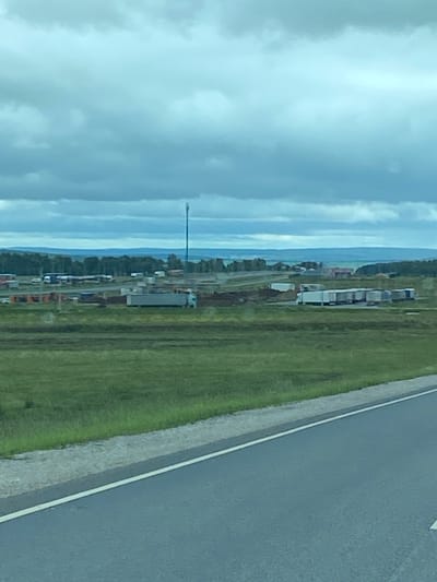 The image depicts an overcast day scene in Kliuchiki, Russia, with a wide-angle perspective from a roadside. In the foreground, a paved road with a white line marking the edge stretches into the distance, bordered by a strip of gravel and then a grassy field. The middle ground reveals an industrial or logistical area with numerous parked semi-trailer trucks and what appear to be shipping containers. Several buildings, some with red roofs, are visible amongst the parked vehicles and mounds of earth or debris. A tall telecommunications tower stands prominently in the background. Beyond this area, a dense line of trees forms a horizon, with rolling hills and a distant body of water under a heavily clouded sky. The overall impression is one of a functional, perhaps rural, transportation hub.