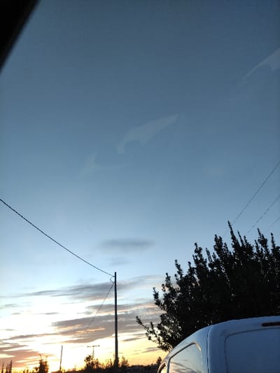 A landscape view at dusk reveals a sky transitioning from blue to shades of yellow and orange near the horizon. A utility pole with wires stretches across the lower right portion of the sky. Silhouetted trees frame the upper right corner, partially obscuring the sky. A reflection appears on a flat surface in the lower left, hinting at a nearby object. The scene presents a serene, rural atmosphere.