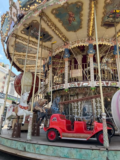The image captures a close-up view of a carousel in Rambouillet, France, likely in a public park given the carousel's intricate design and the presence of surrounding buildings. The carousel is adorned with painted panels depicting cherubs and floral motifs, and illuminated by numerous small lights along its upper edge. Ornate pillars support the canopy, some of which are decorated with carved details.
Several carousel figures are visible: a white horse with red and gold saddle details, a tan dog, and a dark horse with a blue and black saddle. In the foreground, a bright red carousel car, designed like an early automobile with two rows of seating and wooden-effect trim, is prominent. It has red wheels with silver hubcaps. Behind the car, parts of a motorcycle and another carousel vehicle can be seen. A large hot air balloon, red and white, is positioned behind the carousel figures.
A sign on the carousel reads, "LA MAISON NE RE POND PAS DES OBJETS PERDUS OU DETÉRIORES WE ARE NOT RESPONSIBLE FOR LOST OR DAMAGED ITEMS." The background reveals buildings with shutters and awnings, suggesting an urban or semi-urban setting. The sky is blue with some scattered clouds. The carousel appears to be well-maintained and decorated in a traditional style.