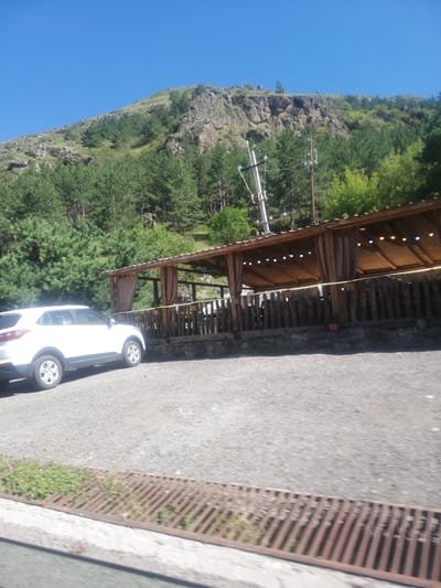 The image shows a white SUV parked on a gravel surface in front of a rustic wooden gazebo with a brown roof. The gazebo is open on the sides, with curtains hanging down, and has lights strung along the underside of the roof. A wooden fence runs along the front of the gazebo. Behind the gazebo, a dense forest of green trees covers the slope of a rocky mountain under a clear blue sky. A metal grate is visible in the lower foreground. The scene appears to be a scenic outdoor dining or resting area, possibly in a mountainous region of Gogasheni, Georgia. There are no people visible in the image.