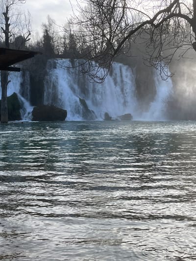 The scene depicts the Kravica Waterfall in Zvirići, Bosnia and Herzegovina. The lower half of the frame is occupied by a body of water with a rippled surface, reflecting diffuse ambient light. Above this, a wide, multi-tiered waterfall cascades over natural rock formations, generating white water and a visible mist plume, particularly on the right side. Dark, moss-covered boulders are present at the base of the falls within the water. The upper background features a dense line of leafless deciduous trees silhouetted against a bright, possibly overcast, sky. A dark, angular wooden structure, likely an overhang, is partially visible in the upper left quadrant, while bare tree branches overhang the upper right portion of the frame. No human figures or animals are present.