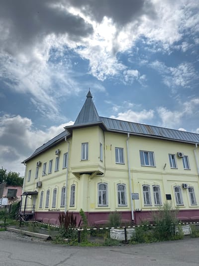 This is a medium-angle, full-frame, eye-level, outdoor, daytime photograph taken in Nogornyi, Russia. The primary subject is a pale yellow, two-story building with a dark gray, conical roofed turret on the left corner. The building has a metal roof and several windows, some with security grates. There are air conditioning units mounted on the exterior walls. The base of the building is a deep maroon color. In front of the building, there is a strip of overgrown grass and plants, with two ornate white planters. A black and white striped barrier runs along the edge of the property. To the left of the main building, a dilapidated pink and gray structure with a broken window is visible. The sky above is a mix of blue and dramatic, cloudy gray formations, with some wispy white clouds. The ground in the foreground is asphalt.