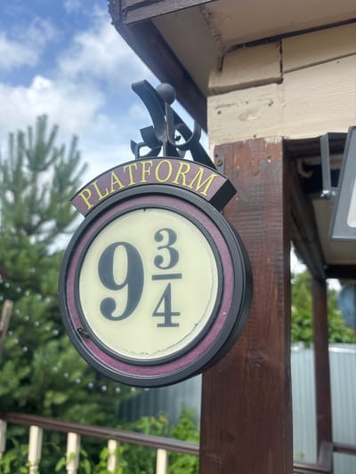 A close-up shot shows a sign for "Platform 9 3/4" hanging from a wooden post. The sign is round with a dark rim and a cream-colored center, featuring the number "9" in large font, and "3/4" in smaller font above and below a horizontal line. The word "PLATFORM" is written in yellow letters on a purple curved piece above the round sign. The background is slightly blurred, showing green trees and a blue sky with white clouds. The wooden post appears to be part of an outdoor structure.