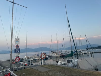 The image depicts a harbor in Gstadt am Chiemsee, Germany, at dusk. Sailboats are moored in the water, with their masts extending towards a pale blue sky tinged with pink and orange hues from the setting sun. In the foreground, a pier juts out into the water, with several signs posted. One sign features a pictogram of a person jumping, with the text "Springen verboten!" (Jumping prohibited!). Another sign indicates "Badestelle" (Bathing area), and a third specifies "Schwimmsteganlage" (Swimming jetty facility) with load-bearing capacity information. A life preserver hangs on a pole. Several people are visible on the pier and on the boats, some appearing to be relaxing or socializing. The water reflects the sky and the boats. In the distance, mountains form a backdrop, their outlines softened by the twilight. The overall atmosphere is peaceful and serene, characteristic of a lakeside town at the end of the day.