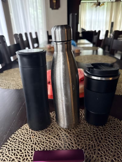 Three reusable drink containers are displayed on a dark wood table. From left to right, they are a black matte tumbler, a brushed stainless steel water bottle, and a black travel mug with a ribbed grip. The containers sit on a light brown, intricately patterned placemat. In the background, several wooden chairs and a glimpse of white curtains are visible. A slice of watermelon is partially visible behind the stainless steel bottle. The scene appears to be an indoor setting.