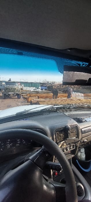 The image displays an outdoor work or storage area in Nazarbek, Uzbekistan, viewed from the interior of a left-hand drive vehicle. The foreground features the vehicle's dashboard, including a black steering wheel with a textured grip, an instrument cluster showing a speedometer and other gauges, and a portion of the control panel on the right side. The base of the windshield and wipers are visible.
The exterior scene is under a clear blue sky, indicating fair weather. The ground surface appears to be dirt or gravel. On the left side, a green, military-style flatbed truck with visible license plates is parked, facing right; one individual is seated or standing on its cargo bed. In the midground, multiple large stacks of sawn timber or wooden planks are centrally located. Three adult individuals are observed in this area: two are positioned among the timber stacks, one wearing dark clothing, and the third is further to the right, also near the materials. Their specific actions involve movement within the work area. In the background, green shipping containers or similar modular structures are visible, alongside other industrial elements and a light-colored van partially obscured.
