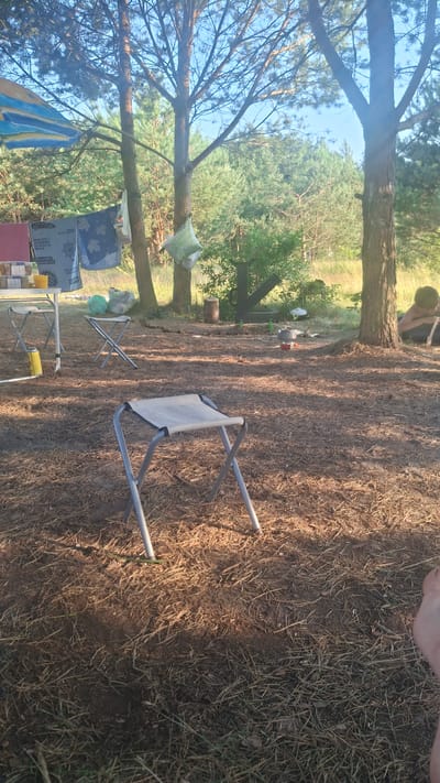 This is a ground-level, medium shot of a campsite scene. The location is identified as Навасвержанскі сельскі Савет, Belarus. The foreground is dominated by dry pine needles and sparse grass on the ground. A light-colored, foldable stool with a metal frame is prominently positioned in the center foreground. In the mid-ground, to the left, a white folding table is visible with a yellow can and some food containers on it. Two other similar foldable stools are placed near the table. A clothesline with laundry is strung between two trees behind the table. To the right of the mid-ground, a person is partially visible sitting near a tree, facing away from the camera. A portable stove with a pot on it is set up on the ground. In the background, there are numerous tall pine trees and green foliage, suggesting a forest environment. The sky is clear blue. Sunlight casts shadows across the scene, indicating it is daytime.