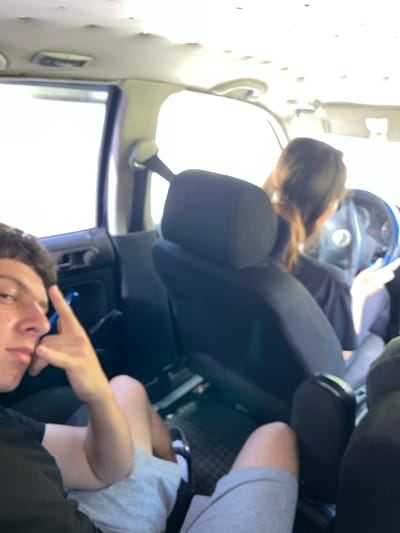 The image depicts the interior of a car, likely in Vlahovo, Bulgaria. Two individuals are visible. In the foreground on the left, a young person with short dark hair is looking towards the camera, resting their chin on their hand and making a peace sign with their other hand. They are wearing a black t-shirt and light grey shorts. To the right and slightly behind, a person with long brown hair, presumably the driver, is looking forward and appears to be holding the steering wheel. They are wearing a black t-shirt. The car's interior shows black seats and a dark dashboard with a steering wheel. The headliner of the car appears to be damaged or sagging in places. The bright exterior light suggests it is daytime.