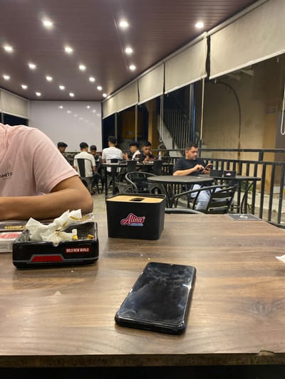 The image presents a nighttime scene at what appears to be an outdoor or semi-outdoor cafe or restaurant in Keudeublang, Indonesia, captured from a first-person perspective. The viewpoint is looking across a wooden table towards other patrons and the surrounding establishment.
In the immediate foreground, a person wearing a light pink t-shirt is partially visible on the left, with text "MMTP" and "Syarifah" faintly discernible on the garment. On the wooden tabletop, a black rectangular container, possibly an ashtray or waste bin, prominently displays "BOLD NEW WORLD" in white and red lettering. Nearby, a smaller black and white box, likely a tissue dispenser, features the brand "Alina" and the phrase "Crunchy Cheese." A black smartphone with a smudged screen lies flat on the table to the right.
Beyond the foreground, several male patrons are seated at dark-colored tables and chairs. One man in a black shirt and light-colored pants is clearly visible, absorbed in his mobile phone. Other individuals are scattered in the background, some partially obscured or facing away, seemingly socializing or relaxing. A black metal railing defines the boundaries of the seating areas.
The setting is illuminated by artificial lights, indicating it is nighttime. A series of small, oval-shaped lights are embedded in a reddish-brown wall on the far left, contributing to the ambient glow. White awning-like structures are visible above the seating areas. A small black sign with a white "P" and "6" can be seen on a table in the background. The overall atmosphere is casual and relaxed, with patrons engaged in their own activities.