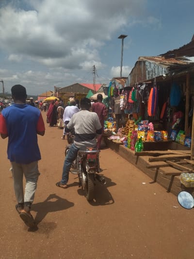 The image shows a street scene in Lokoja, Nigeria, taken at approximately 7.404 latitude and 7.632 longitude. It is a dusty, dirt road lined with shops and vendors. The shops appear to be made of corrugated metal and wood. The vendor's goods are displayed on makeshift stalls. The scene is bustling with people and activity. Several people are walking along the street, while others are shopping or engaging with the vendors. There is a man riding a motorcycle toward the camera. There is also a streetlamp in the background and a round mirror on the ground in front of the shops. The sky is clear and partly cloudy.