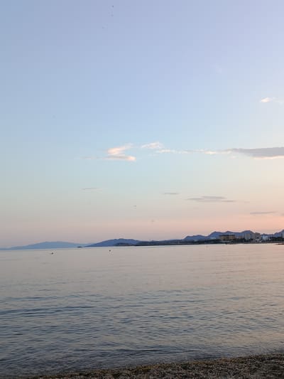 The image captures a serene coastal scene in Águilas, Spain. The sky is a gradient of light blue and soft pink hues, typical of sunset or sunrise. A few wispy clouds are scattered across the sky. The calm water of the Mediterranean Sea stretches out, reflecting the colors of the sky. In the distance, a coastline of mountains and buildings is visible. The foreground consists of a pebble beach. There are a few small yellow buoys floating on the water. Several small objects are visible in the sky, possibly birds or other airborne items.