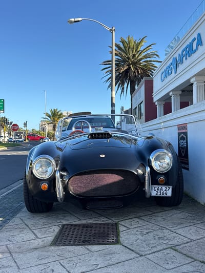 A low-angle, head-on shot captures a black convertible sports car, identified by its license plate as "CAA 27 2164," parked on a paved sidewalk. The car's design features prominent round headlights, a large meshed grille with a reddish interior, and a sleek, curved hood with an air intake scoop. A clear windshield and a roll bar are visible. The car is positioned in front of a white building with architectural columns and the blue lettering "CARS 4 AFRICA" above it. To the left of the car, a street with traffic lights and a stop sign is visible, along with other vehicles and palm trees. The scene appears to be in Blouberg, South Africa, indicated by the "CARS 4 AFRICA" establishment. The sky is clear and blue.
