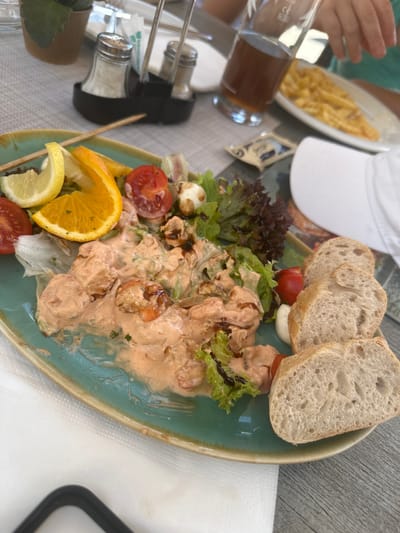 The image shows a close-up of a meal served on a teal-colored plate. The plate contains a salad with lettuce, tomatoes, and small white balls that appear to be mozzarella. There are also several slices of bread and a creamy pink sauce with visible pieces of shrimp. A slice of orange and a wedge of lemon are also on the plate. The meal is set on a wooden table, with a white napkin visible in the foreground. In the background, a salt and pepper shaker set is on a table, as well as a glass of dark liquid and a plate of french fries. The setting appears to be an outdoor dining area, possibly a restaurant or cafe. The lighting suggests it is daytime. There is no visible indication of the specific location within Schönau am Königssee, Germany, beyond it being a place where food is served.