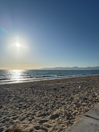 The image depicts a beach scene in Les Meravelles, Spain, at sunrise or sunset. The sky transitions from a deep blue to a hazy yellow as the sun approaches the horizon. A faint contrail from an airplane is visible high in the sky. The ocean is calm with small waves reflecting sunlight, creating a shimmering effect. The beach is sandy and covered in footprints, indicating recent human activity. A concrete sidewalk or pathway runs along the edge of the beach.