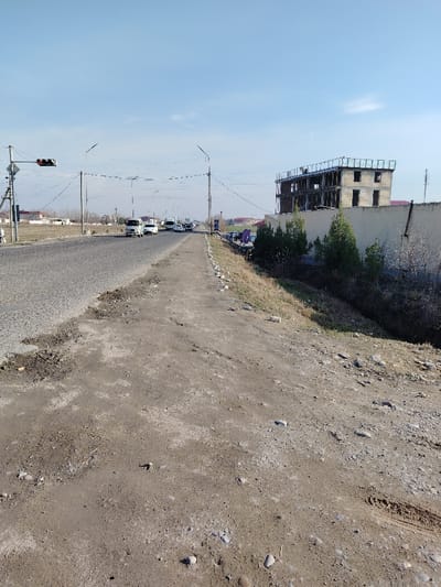 The image depicts an exterior view of a road and its immediate surroundings in Juma, Uzbekistan, under a clear blue sky. The foreground is dominated by an unpaved, uneven dirt and gravel path with scattered small rocks and visible tire imprints on the right. To the left, an asphalt two-lane road extends into the distance, showing signs of wear and patching. A traffic signal displaying a red light is visible on the left side of the road, mounted on a utility pole. Multiple utility poles with overhead wires span across and alongside the road.
In the mid-distance on the road, several white vehicles, including what appear to be vans and cars, are traveling away from the viewer. To the right of the road, a strip of sparse vegetation and dirt slopes down into a narrow, dry ditch. Beyond the ditch, a light-colored solid wall runs parallel to the road. Behind this wall, on the right side of the frame, a multi-story building, approximately three to four stories tall, is under construction. It exhibits an unfinished concrete frame structure with exposed brickwork and no visible windows or exterior finishes, topped with a skeleton roof structure. Green conical evergreen trees and other sparse dry vegetation are present along the wall and in the ditch area. Distant buildings and a faint industrial silhouette are visible on the horizon. No individuals are visible in the scene.