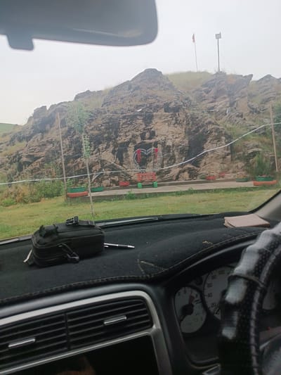 An indoor, dashboard view captures an outdoor scene through a car windshield. The exterior shows a large, rocky mountainside behind a grassy area with a decorative heart-shaped wreath on a bench, positioned in front of the rock face. Several potted plants are visible in front of the bench. A flag flies from a pole on the crest of the hill. Inside the car, the dashboard is cluttered with a black pouch and a pen. The steering wheel and part of the instrument cluster are also visible on the right side of the image. The overall lighting suggests an overcast day.