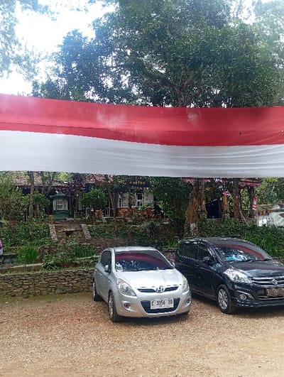 This image depicts an outdoor scene in Kajar, Indonesia, featuring a parking area and a multi-level structure set into a verdant hillside.

In the immediate foreground, a light brown gravel or dirt surface serves as a parking area. Two passenger vehicles are parked side-by-side: on the left, a silver Hyundai i20 with Indonesian license plate "E 3356 DD," and on the right, a black Suzuki Ertiga with Indonesian license plate "B 1951 PK." Both vehicles have sunshades visible through their windshields.

Above the cars, a large, horizontally stretched banner, red on top and white on the bottom, spans across the frame, partially obscuring the view of the background. This color scheme resembles the Indonesian national flag.

Behind the banner and parked vehicles, the terrain rises, supported by terraced stone retaining walls. These walls and the elevated ground are densely covered with green foliage, including various plants and shrubs. Further back and higher on the incline, multiple buildings are visible, partially obscured by the banner and trees. One building section has a sign that reads "coffee" in white letters. The architecture suggests a casual, possibly commercial or recreational complex, integrated with the natural landscape.

The upper portion of the image is dominated by the dense green canopy of large, mature trees, set against a bright, overcast sky. No people are visible in the frame. The scene suggests a daytime setting, possibly a cafe or recreational spot.