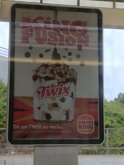 This is a photograph of a Burger King advertisement for a King Fusion Twix dessert, displayed on an illuminated billboard in Seixal, Portugal. The billboard, measuring approximately 2.5 meters tall and 1.5 meters wide, is mounted on a concrete pedestal. The advertisement features a close-up of the King Fusion dessert, a layered ice cream sundae with Twix candy, chocolate sauce, and cookie pieces, served in a clear glass. The Burger King logo is prominently displayed in the lower right corner. The text on the advertisement is in Portuguese, promoting the dessert as a summer treat. The background of the advertisement shows a stylized red and yellow sunburst pattern. The billboard is positioned outdoors, with a blurred background of lush green trees and a cloudy sky. In the lower foreground, a metal railing and a paved walkway are visible, suggesting a public area like a park or promenade. The image is taken from a slightly angled perspective, with reflections visible on the billboard's surface.