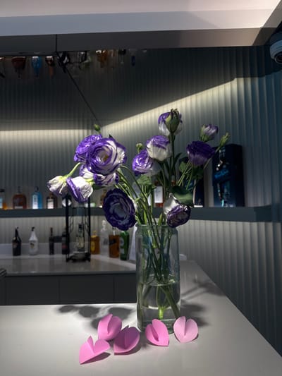 A vase with blue and white flowers sits on a white surface next to pink paper cutouts. The background features a modern interior with horizontal blinds and a shelf stocked with various bottles, possibly liquor. The lighting is bright, casting shadows and highlighting the textures of the flowers and paper cutouts. The scene is simple, yet the color contrast and lighting create a visually appealing composition.