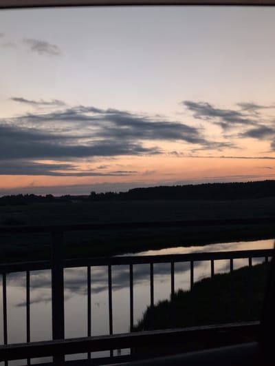 This image depicts a serene natural landscape during twilight, viewed from what appears to be a bridge or elevated walkway. The sky is a gradient of soft blues, grays, and oranges, with layered clouds stretching across the horizon. Below the sky lies a dark silhouette of trees and fields, suggesting a rural or parkland setting. In the foreground, a dark railing with vertical bars is visible, separating the viewer from a body of water that reflects the muted colors of the sky and clouds. The overall impression is one of calm and natural beauty at the close of day. The location is specified as Trubichiino, Russia.