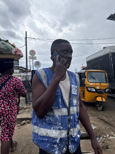 The image shows a medium shot of a Black man on a phone call, wearing a blue and white reflective vest. He is standing on a dirt and paved street, possibly in Lagos, Nigeria. Behind him, a yellow auto-rickshaw, commonly known as a "Keke," is parked. The man's vest has some paint splatters on it.
In the foreground, another person is partially visible, carrying a load of produce on their head, and wearing a patterned dress. The scene appears to be a busy street market or roadside area. Several signs are visible in the background, including one with circular logos and text that appears to say "Ozé's" and "dz scentz." Another sign reads "HIC PARLON Salon".
The sky is overcast, suggesting a cloudy or potentially rainy day. The overall impression is that of a typical day in a bustling urban environment in Nigeria, with people engaged in commerce and daily activities. The man's focus on his phone call indicates he is engaged in a conversation, possibly for work or personal reasons, while navigating the busy surroundings.
