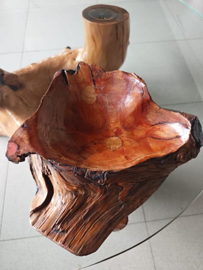A carved wooden bowl with a rich reddish-brown hue sits on a glass table. The bowl's interior displays concentric growth rings and is polished to a high sheen, reflecting light. Three circular inlays are visible within the bowl's surface. The exterior of the bowl features a rough, gnarled texture with dark, charred areas and intricate wood grain. Behind the bowl, a section of a larger wooden structure, possibly a table leg or base, is visible. The floor appears to be tiled in a light, neutral color. The overall scene is likely an indoor setting, possibly a home or workshop.