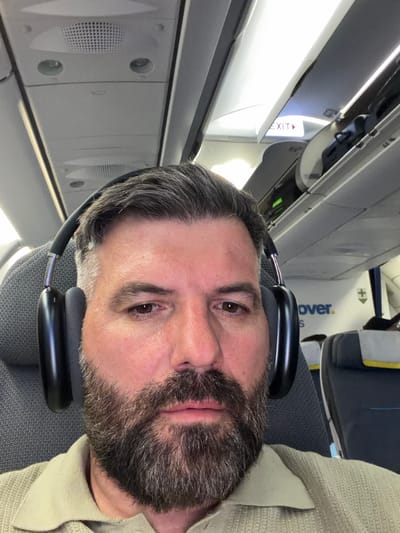 A man is in the cabin of an airplane, wearing black headphones. The cabin interior is visible, including the overhead compartment with an 'EXIT' sign, individual air vents, and a luggage storage compartment. The airline's logo is partly visible on the overhead compartment door above the seating. The man has grey hair and a salt-and-pepper beard and is wearing a beige knitted garment. The photograph appears to have been taken by the man himself. The location of Paradise, United States of America, is not detectable within the image.