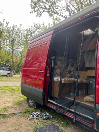 The image depicts a red van with its side door open, parked on a grassy area. A mesh screen is in place at the doorway. Inside the van, elements of a living space are visible, including what appears to be a sleeping area and storage. The setting is outdoors, with trees and foliage in the background, suggesting a natural environment. A mat with the word "Bearbrick" is outside the door along with two pair of sneakers. A bicycle and white vehicle can be seen further in the background beyond the trees. The specific location is stated to be Kastraki, Greece.