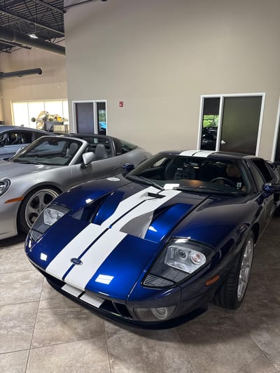 The image shows two Ford GT cars parked indoors, likely in a showroom or private garage in West Chicago, United States. The car in the foreground is a dark blue Ford GT with white racing stripes, while the car behind it is a silver Ford GT. The cars are parked on a tiled floor, and the background shows neutral-colored walls with windows reflecting outdoor scenery. Fluorescent lighting is present. There are no people immediately visible.