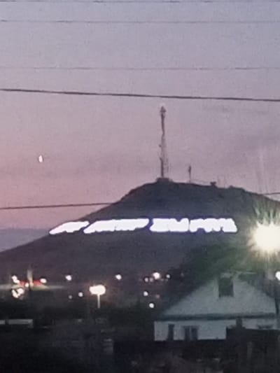 The image shows a hillside with white illuminated lettering on it. The lettering appears to spell out "ҚАЛАШЫҚ" which translates to "city" or "settlement". At the top of the hill, there is a telecommunication tower. In the foreground, there is a building with a white facade and a dark roof, possibly a house or a smaller structure. Several streetlights are visible in the foreground and around the base of the hill. The sky is dusky purple, suggesting it is either dawn or dusk. There is a small, bright object in the sky to the left of the hill, which could be a planet or a distant light. The scene is likely located in Botakara, Kazakhstan, given the presence of the lettering and the landscape.