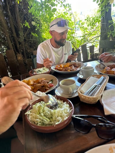 The image depicts a meal being enjoyed outdoors, likely at a restaurant or cafe in Dulovine, Montenegro. A man wearing a white Fila t-shirt and a white cap with purple-tinted goggles is seated at a wooden table. He is looking at his phone and holding a fork in his right hand, appearing to be eating. Several plates of food are visible on the table, including a dish with meat and potatoes, a bowl of what appears to be shredded cabbage salad, and other partially eaten meals. There are also cups of coffee and a basket containing cutlery. The background shows lush green foliage and trees, suggesting a natural or garden setting. The overall scene conveys a casual dining experience.