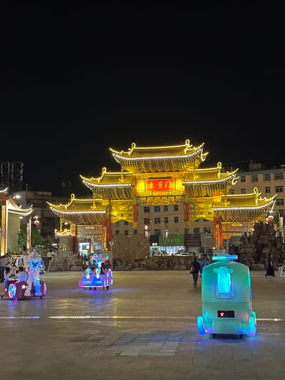 The image captures a nighttime scene in Eshan Yi Autonomous County, China, likely a public square or pedestrian area. A large, ornate, multi-tiered gateway, illuminated by golden lights, dominates the background. The gateway features traditional Chinese architectural elements like curved eaves and decorative carvings. In front of the gateway, on the paved ground, are several small, battery-operated children's rides.

In the mid-ground, two of these rides are prominent. One on the left is a red car-shaped ride with a large robot figure at the front, carrying at least one child. Another ride to its right is a colorful, cartoonish vehicle with several children seated inside. Both rides are lit with vibrant LED lights in blue, pink, and green. Further to the right, another light-green ride, shaped like a futuristic pod, is also illuminated and parked.

Several people are visible in the scene, mostly walking or standing. A few individuals are seen near the rides, presumably supervising or waiting. A group of people is walking away from the camera in the mid-ground, towards the gateway. In the far background, beyond the gateway, buildings are visible, also lit up. The overall atmosphere suggests a lively evening in a public space that caters to families and entertainment. The ground surface appears to be paved, with some areas showing a polished or reflective quality due to the lighting. The night sky is dark, with no stars or moon visible.