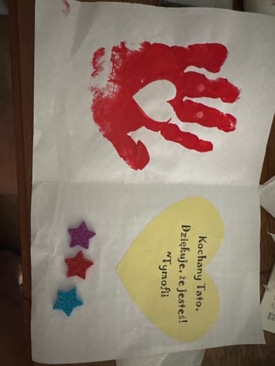 The image shows a handmade card. The top of the card features a red handprint with a white heart shape in the center of the palm. Below the handprint, there are three glittery star stickers arranged vertically: purple, red, and blue. To the right of the stars is a yellow heart-shaped cutout with black writing in Polish. The writing says "Kochany Tato, Dziękuję, że jesteś! ^ I'm ofi" which translates to "Dear Dad, Thank you for being you! ^ I'm ofi". The card is on a white piece of paper, which is placed on a wooden surface. The overall scene appears to be indoors, possibly at a home. There is no visible information in the image that would allow for a specific location in Sosnowiec, Poland.