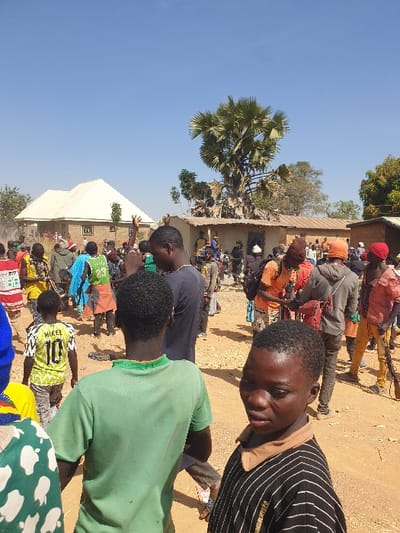 A crowd gathers outdoors near a building. Many people are seated or standing. A young boy in a striped shirt appears in the foreground. Behind him, other children wear brightly colored clothing. Palm trees and other vegetation are visible. The scene is brightly lit under a clear blue sky. The composition is shot from a low angle, emphasizing the height of the crowd and the surrounding environment.