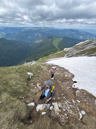Two individuals are on a mountainous terrain near Jošanica, Montenegro. The person in the foreground, wearing a grey and black patterned cap, a blue t-shirt, and a black and yellow plaid jacket, is raising their right hand with their palm facing forward, appearing to gesture or pose. They are positioned on a grassy slope with scattered rocks. Behind this individual, a second person, wearing a pink jacket and black pants, is taking a photograph with a pink camera. This person is situated on a rocky path adjacent to a patch of snow. The background reveals a vast expanse of rolling, forested mountains under a cloudy sky, characteristic of the mountainous landscape in the region of Jošanica.