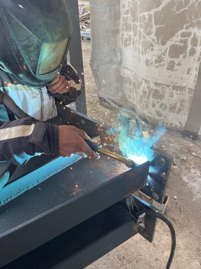 The image shows an individual engaged in arc welding an industrial metal structure outdoors in Zlatovrah, Bulgaria. The welder, positioned from the waist up, is wearing a dark protective welding helmet with a tinted visor, a light-colored zippered jacket, and dark long-sleeved gloves with light stripes. The individual holds a welding torch with a brass-colored tip and black handle, generating a bright blue-green arc and numerous orange sparks as metal components are joined. The subject of the welding is a structure composed of several dark-painted, heavy-gauge steel beams, with visible bolt holes and existing welds. A black cable, likely a ground connection, is clamped to the metal structure. The immediate background to the right features a large, light-colored fabric or tarp with a cracked, mottled texture, potentially serving as a screen or barrier. Further in the background, out-of-focus stacked materials, possibly wood or construction debris, are visible, suggesting an industrial or construction site. The ground appears to be a concrete or paved surface.