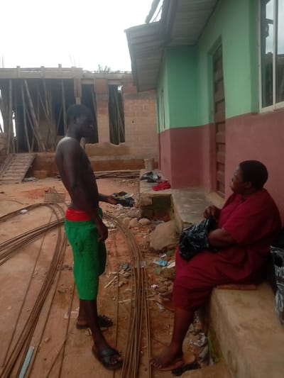 In Ihiala, Nigeria, an outdoor scene depicts a shirtless, dark-skinned man wearing green shorts with red trim and black sandals, standing on a dirt ground with reinforcing bars. He is holding a dark object in his right hand. Beside him, a woman in a red dress sits on a concrete bench. She has a black plastic bag on her lap. They appear to be in conversation. Behind them, a building is painted green and red. In the background, there is a partially constructed building with bamboo supports. The ground is littered with debris.