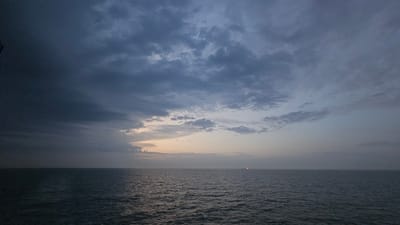 This image depicts a wide, dark seascape under a heavily clouded sky, likely taken at dusk or dawn. The sea is a deep, textured blue-grey, with gentle ripples indicating a calm to moderate water condition. The horizon is a stark, straight line dividing the sea from the sky. The sky is filled with dramatic, dark grey clouds that dominate the upper portion of the frame. Lighter patches of sky are visible towards the center-right, where the sun appears to be setting or rising, casting a faint, warm glow. A tiny, distant red light, possibly from a vessel or a marker, is visible on the horizon to the right. The overall impression is one of a vast, slightly melancholic natural environment.
While the image is of a seascape, it is impossible to determine the specific location in Dover, United Kingdom, based solely on the visual information provided. Dover is a coastal town, and this image could have been taken from its coastline or from a vessel offshore. However, without any landmarks or identifiable features, pinpointing a precise location within Dover is not feasible.